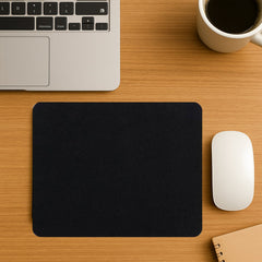 A black mouse pad on a wooden desk with a white mouse, laptop, coffee cup, and notebook.