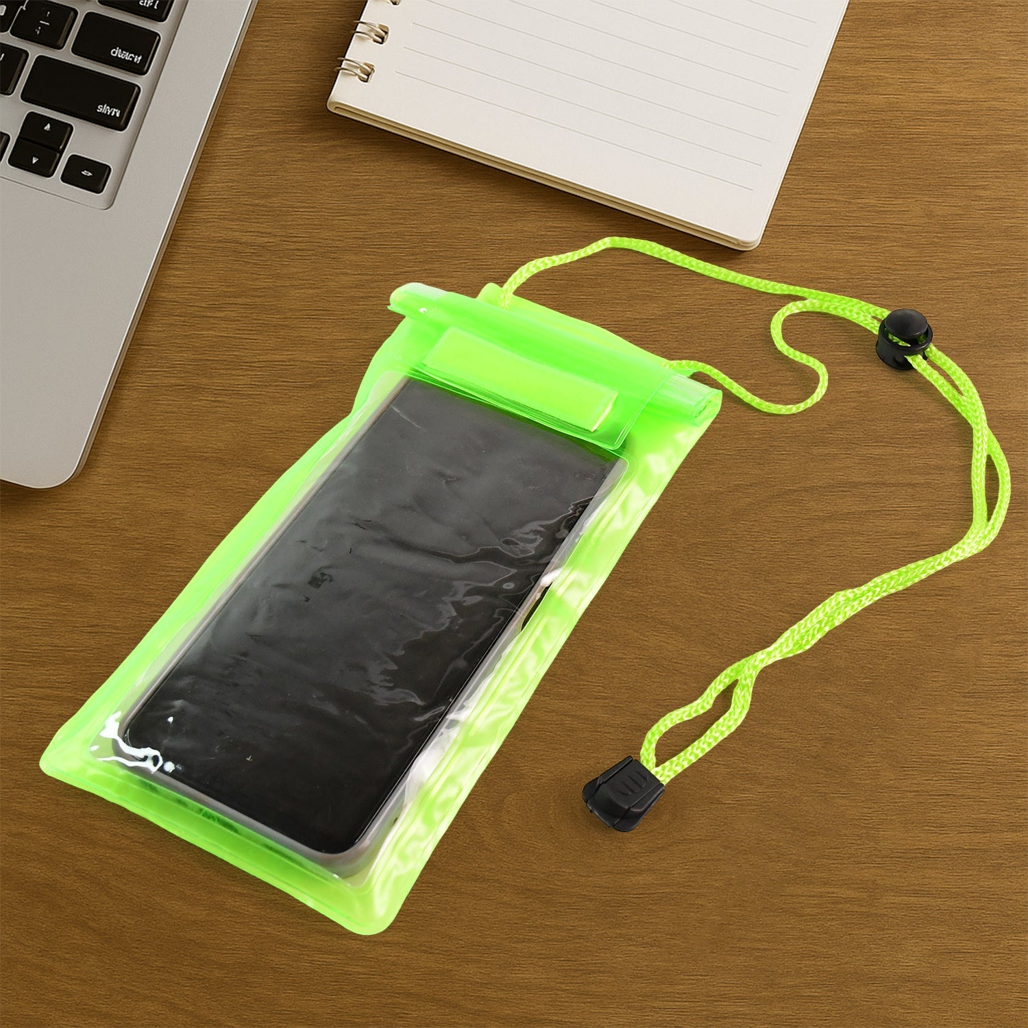 Neon green waterproof phone pouch with a black phone inside, lying on a wooden desk next to a laptop and notebook.