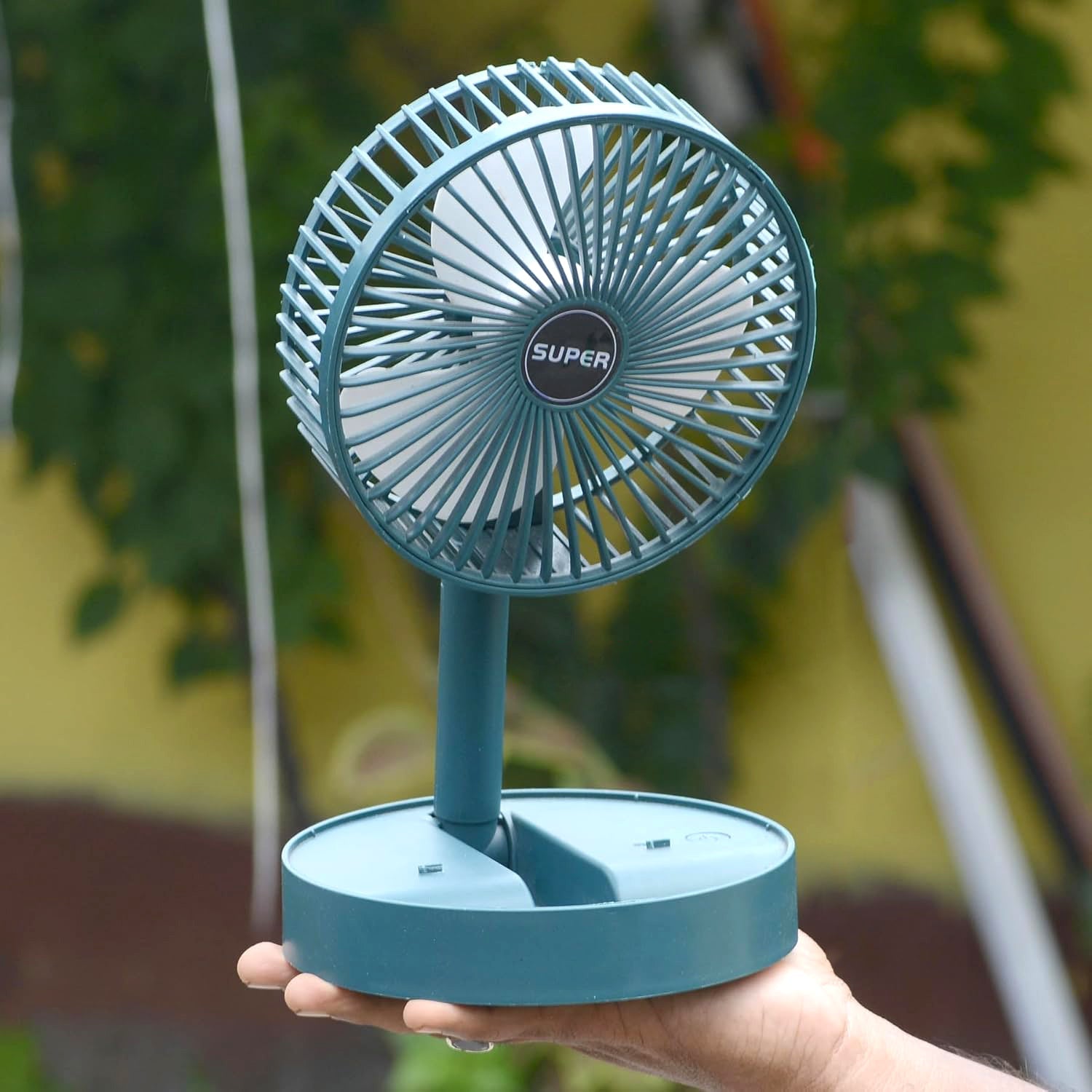 A hand holds a teal portable fan with an adjustable stand, against a blurred outdoor background.