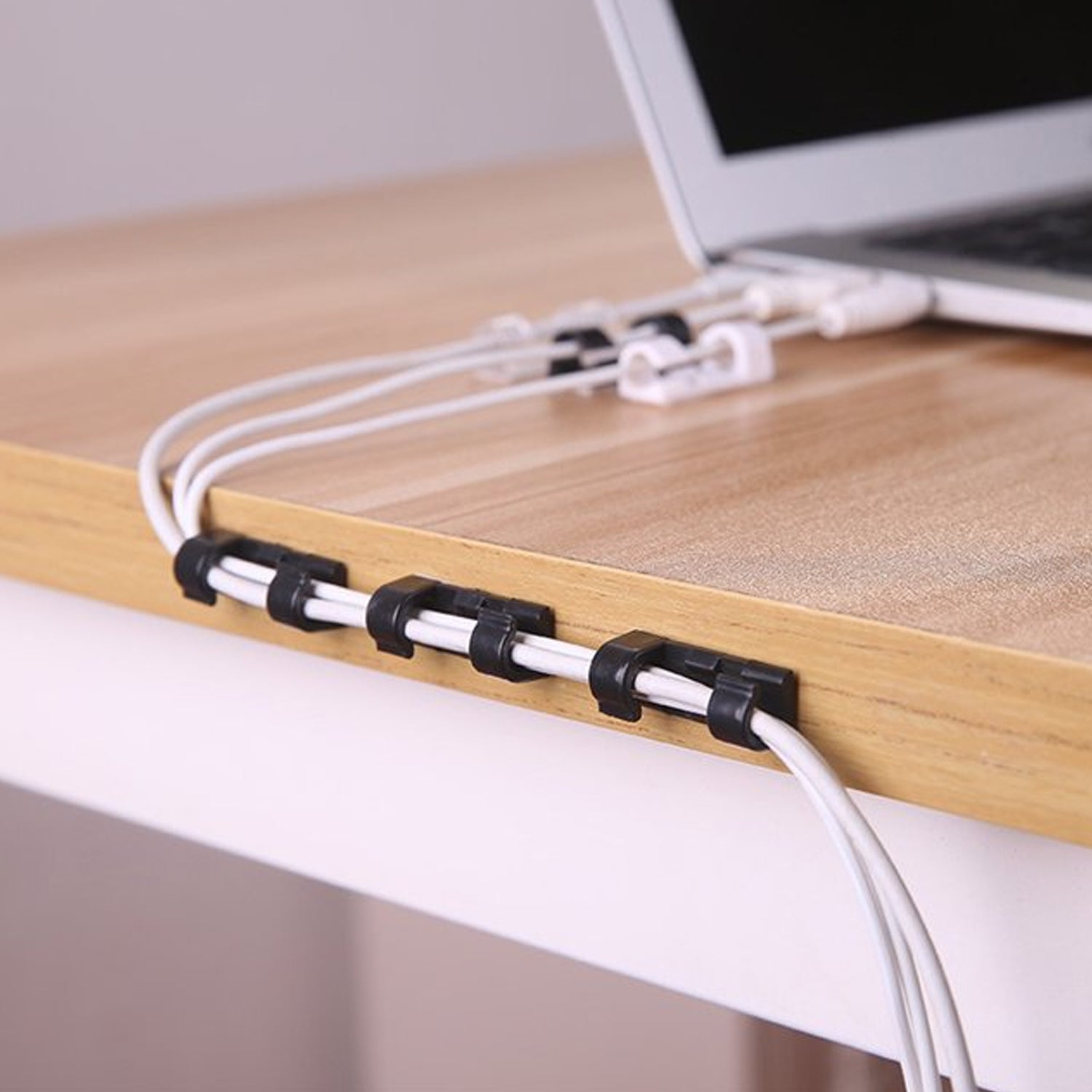 Three black adhesive cable clips securing white cables to the side of a wooden desk, with a laptop in the background.
