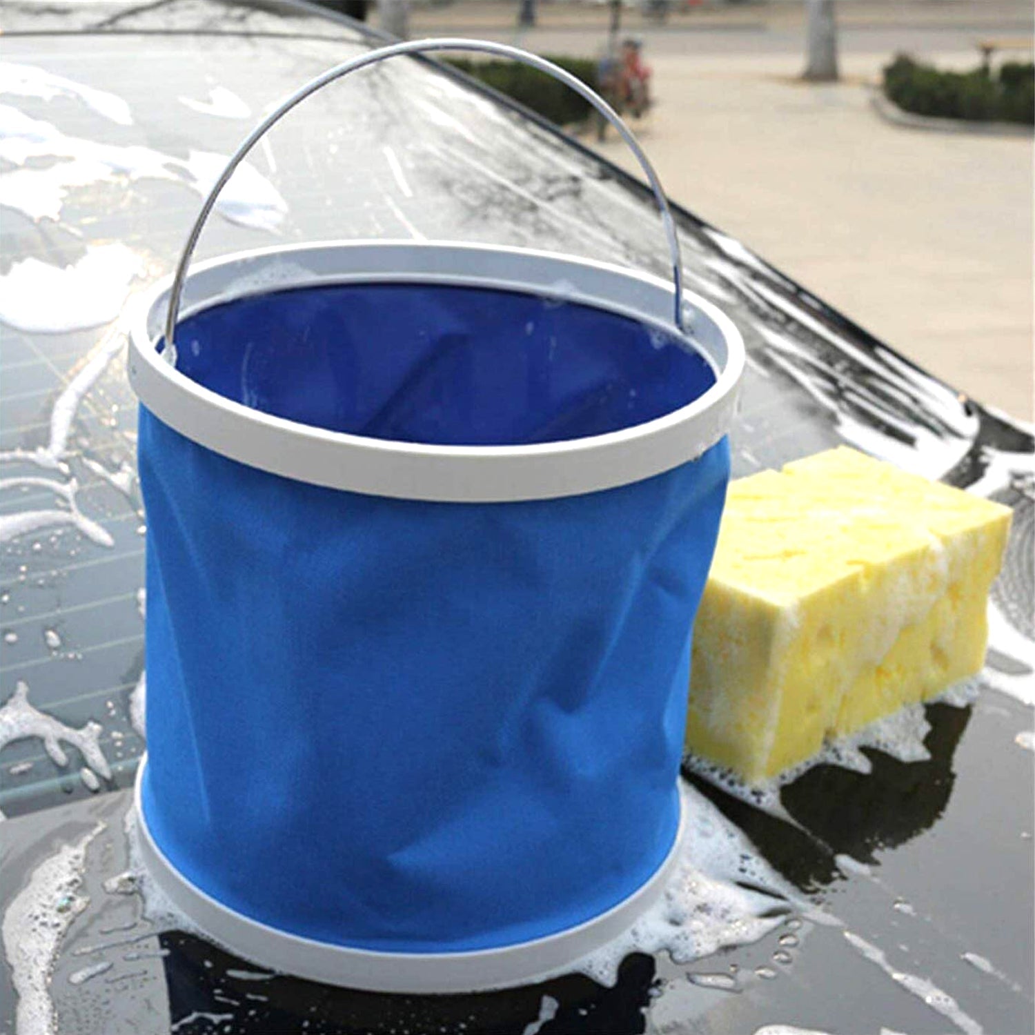 A blue collapsible bucket and a yellow sponge on the soapy roof of a car.