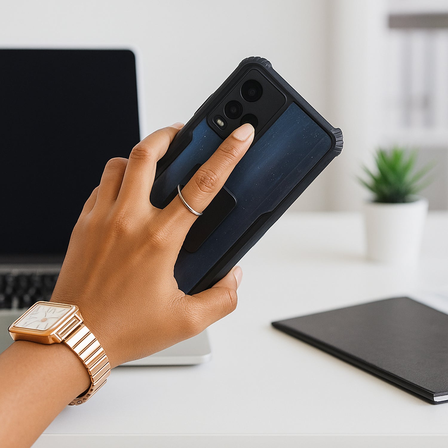 A hand holding a black smartphone with a silver ring stand, with a laptop and notebook in the background.