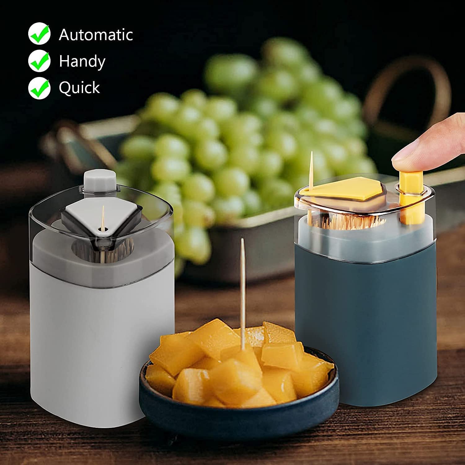 Two automatic toothpick dispensers, one grey and one teal, on a wooden table with a bowl of mango cubes and grapes in the