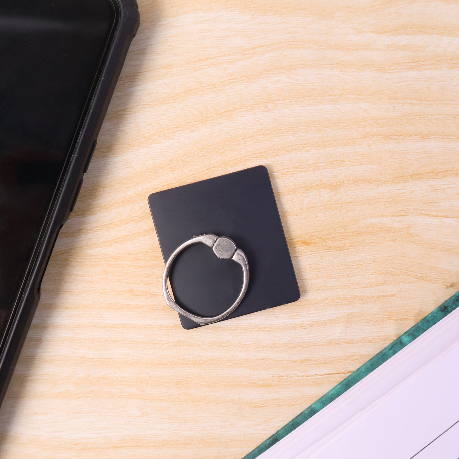 A black square phone ring stand with a silver ring next to a phone and an open book on a wooden table.