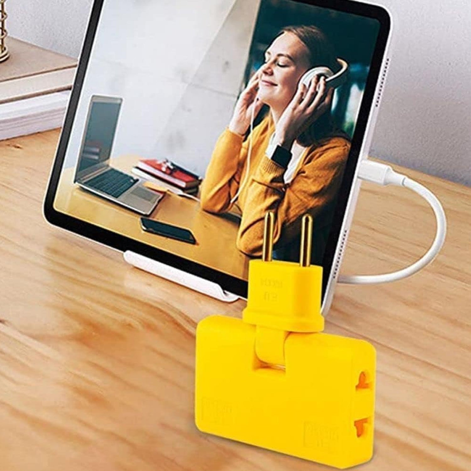 A yellow 3-in-1 foldable wall plug adapter on a wooden desk, with a tablet displaying a woman wearing headphones.