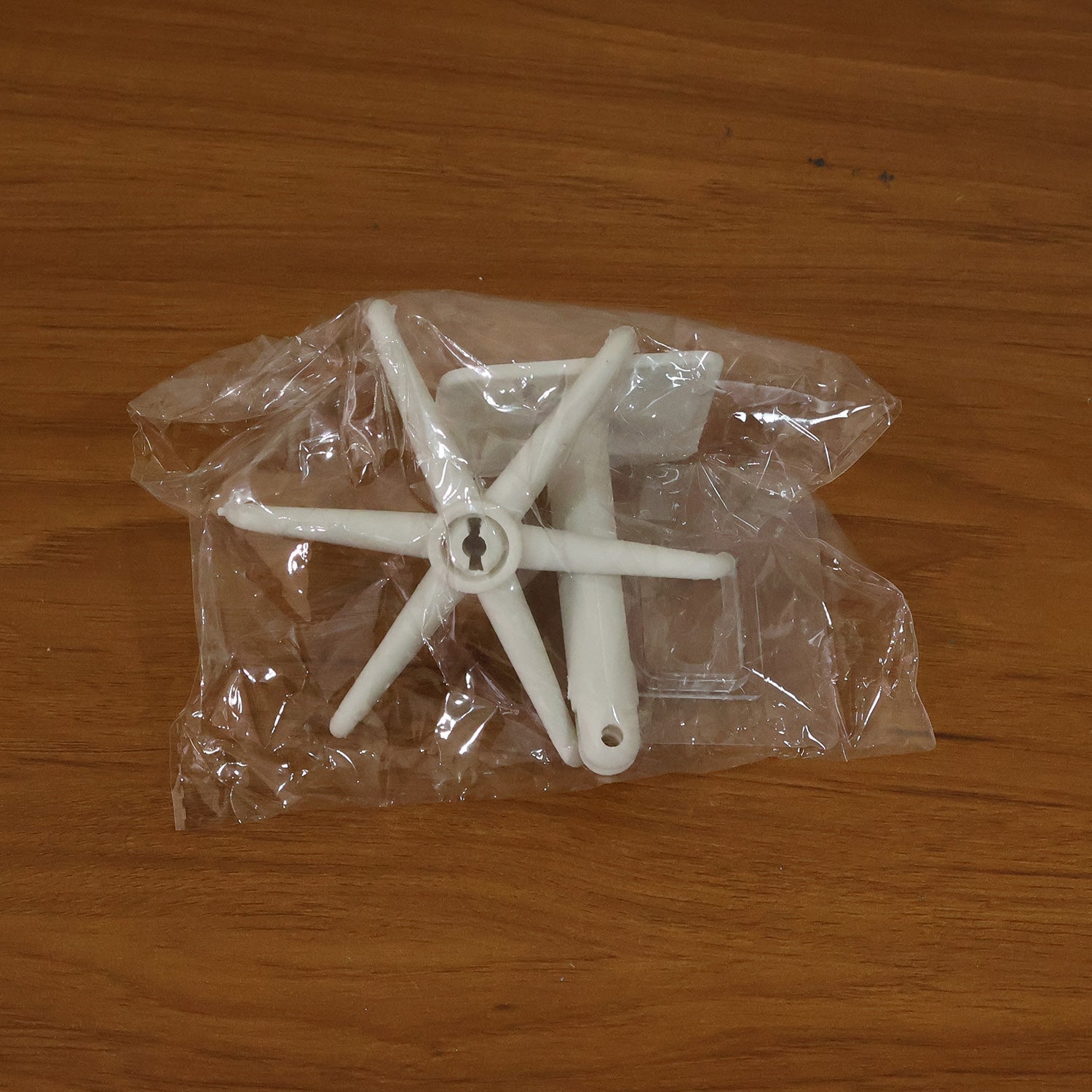The white plastic components of the rotating hook are sealed in a clear plastic bag on a wooden surface.