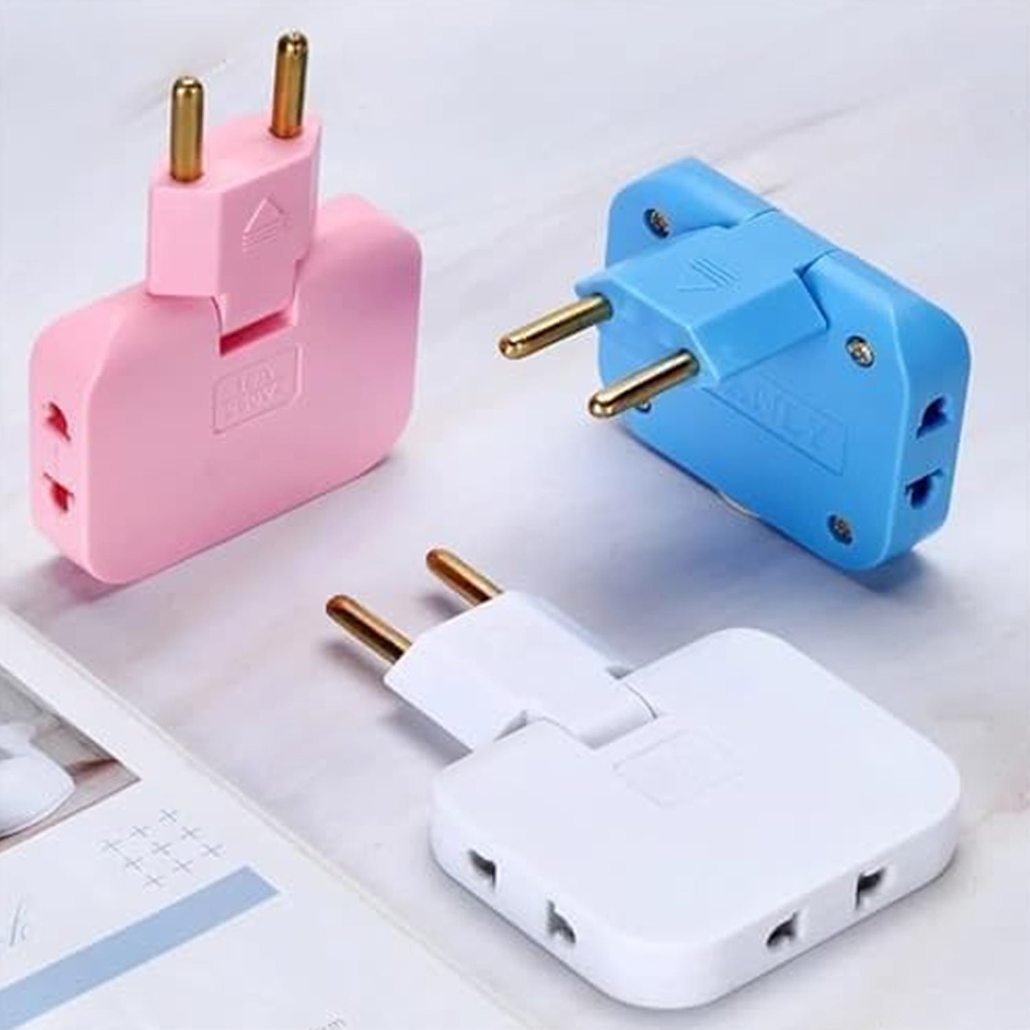 Three 3-in-1 foldable wall plug adapters in pink, blue, and white, arranged on a light surface.