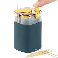 A hand presses the yellow button of a teal automatic toothpick dispenser, releasing a single toothpick. Several toothpicks