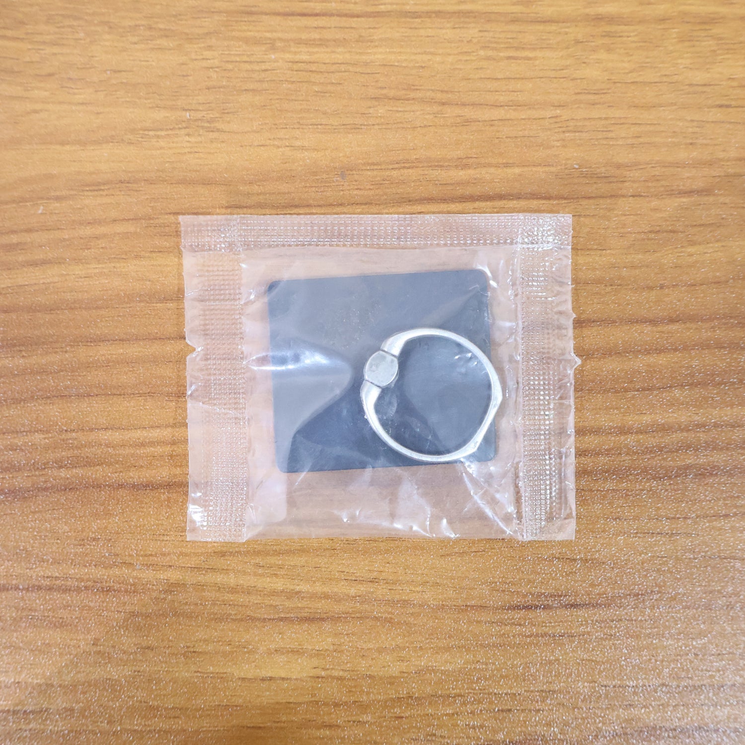 A black square phone ring stand with a silver ring, sealed in clear plastic packaging, on a wooden surface.