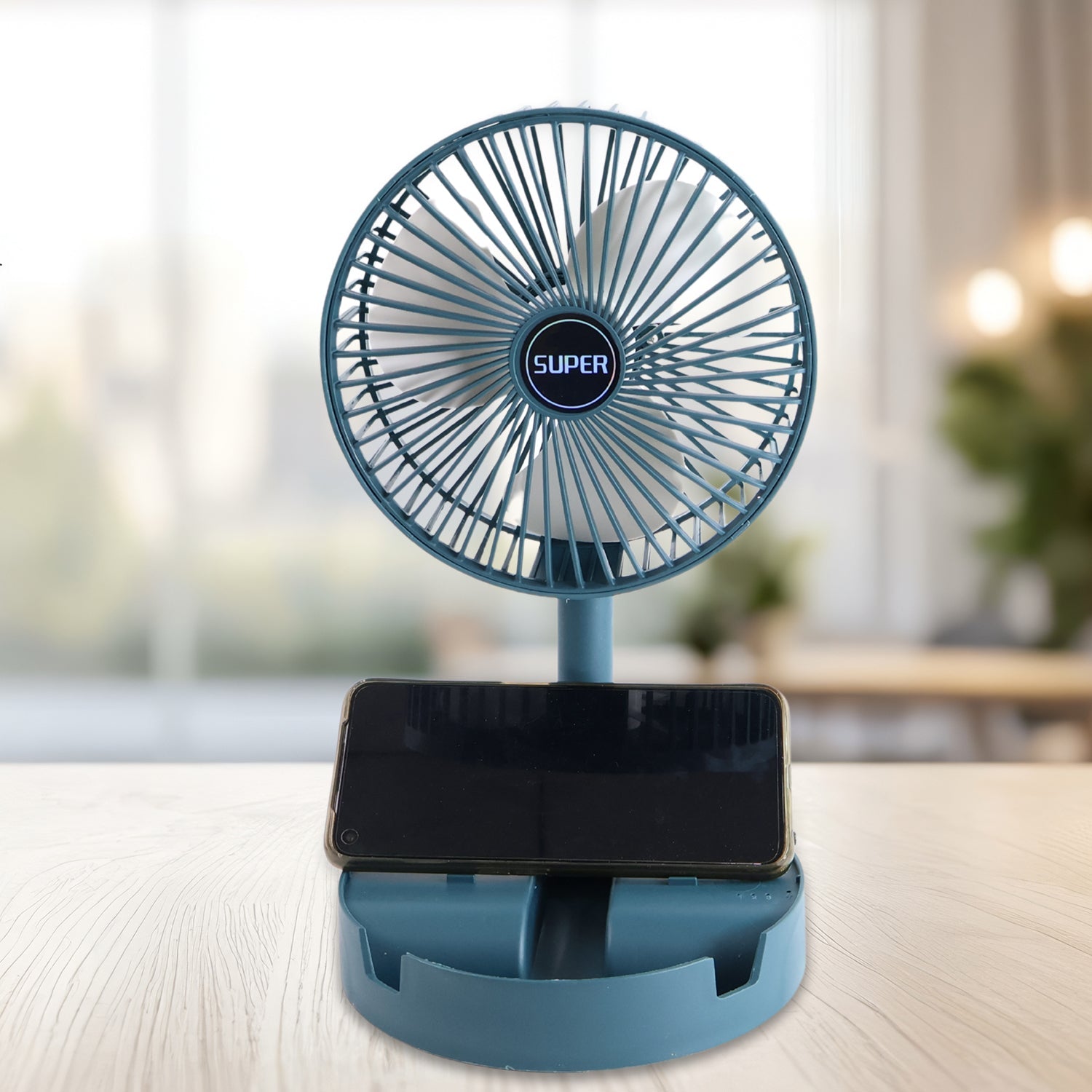 A teal portable fan with a phone resting in its base is on a wooden table, with a bright window in the background.