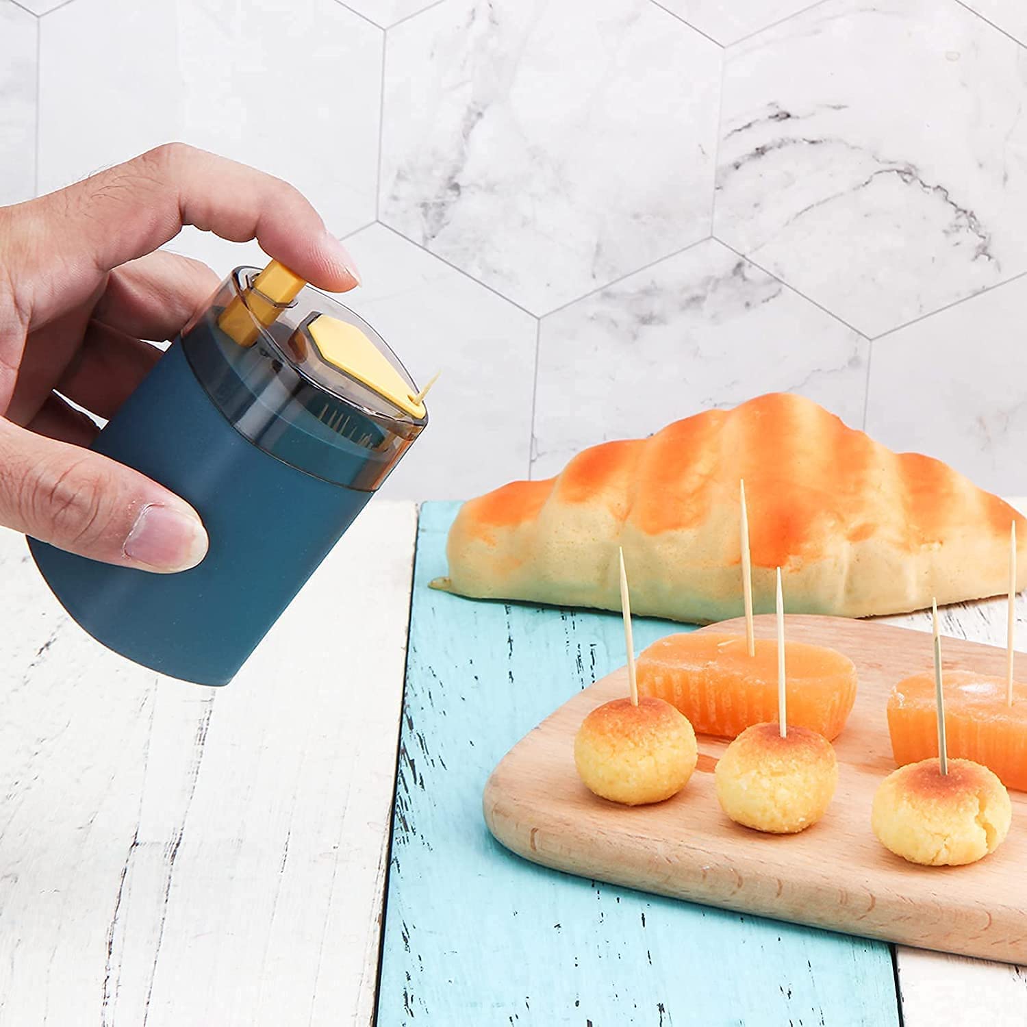 A hand holds a teal automatic toothpick dispenser, next to a wooden board with small food items and a croissant in the