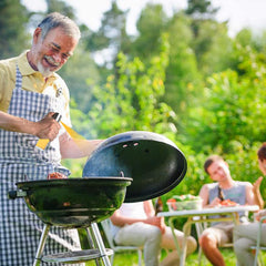 A smiling man in a checkered apron sprays a barbecue grill with an oil sprayer, with people in the background.
