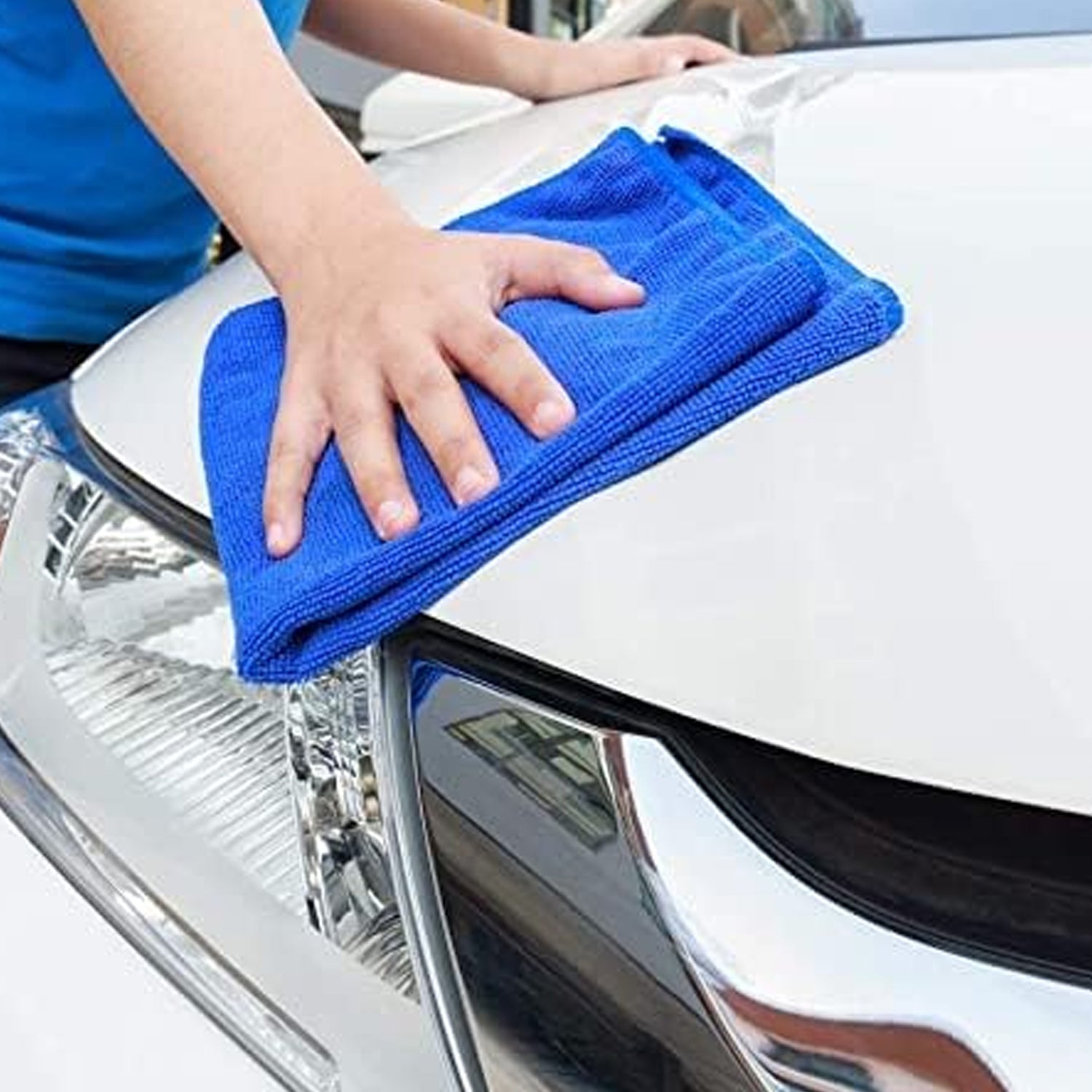 A hand in a blue shirt wiping the hood of a white car with a blue microfiber cloth.