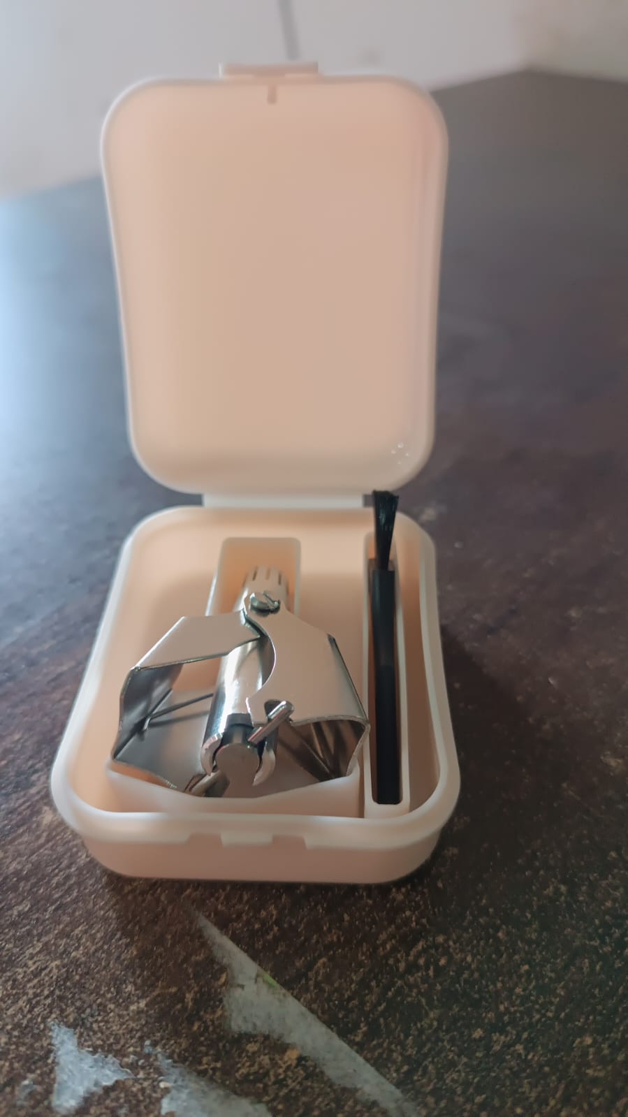 A silver manual nose hair trimmer and black brush are neatly packed inside a small open beige plastic case.