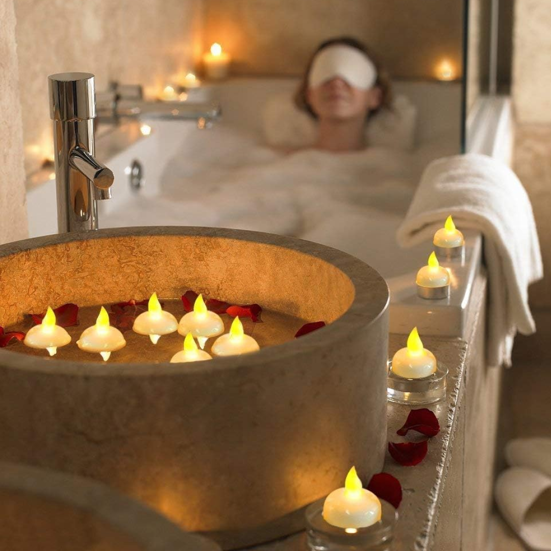 A stone basin filled with water, red petals, and white LED floating candles, with a woman bathing in the background.