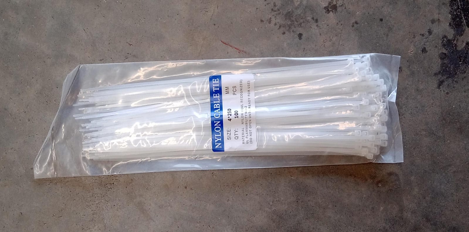 A clear plastic package of white nylon cable ties with a blue label, lying on a concrete surface.