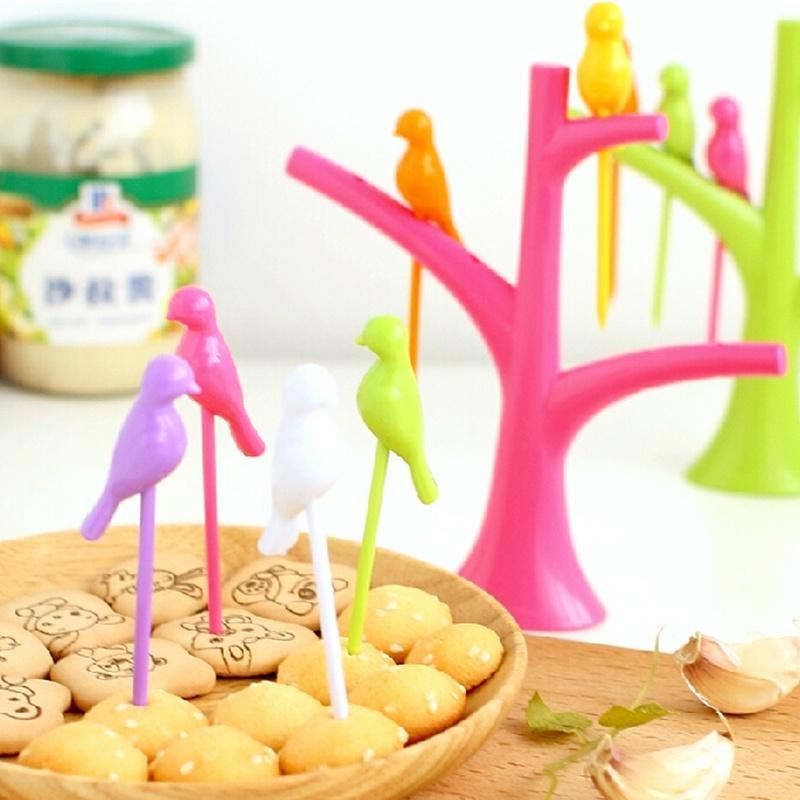 A pink tree holder with colorful bird fruit forks in a bowl of cookies, with a green tree holder and jar in the background.