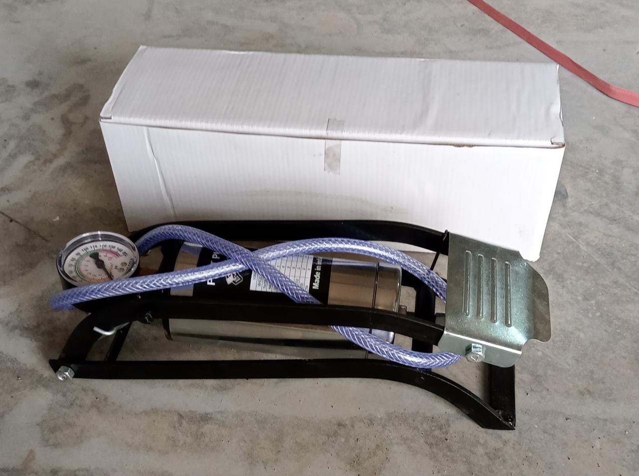 A black and silver foot pump with a hose and gauge is placed in front of a white cardboard box on a concrete floor.