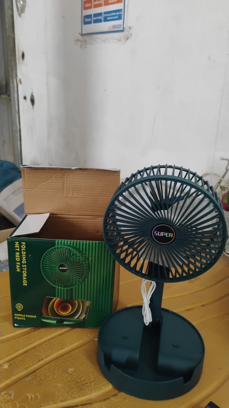 A teal portable fan with its green and black packaging box is on a textured yellow surface.