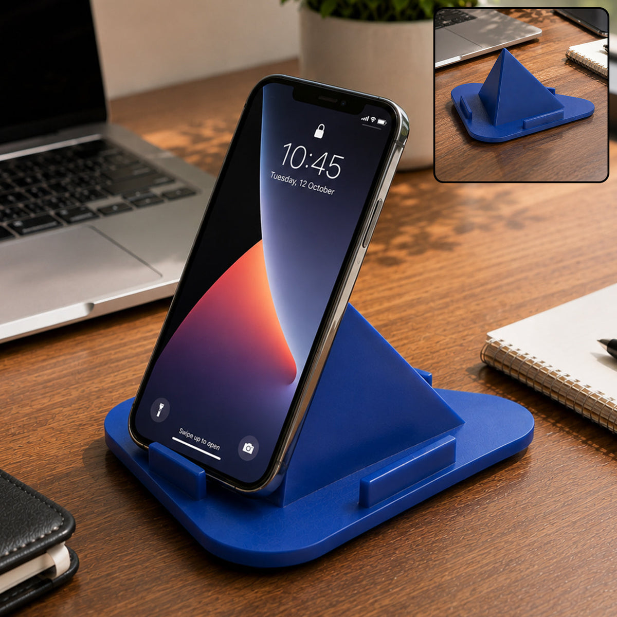 A blue pyramid mobile stand holds an iPhone on a wooden desk with a laptop and notebook nearby.