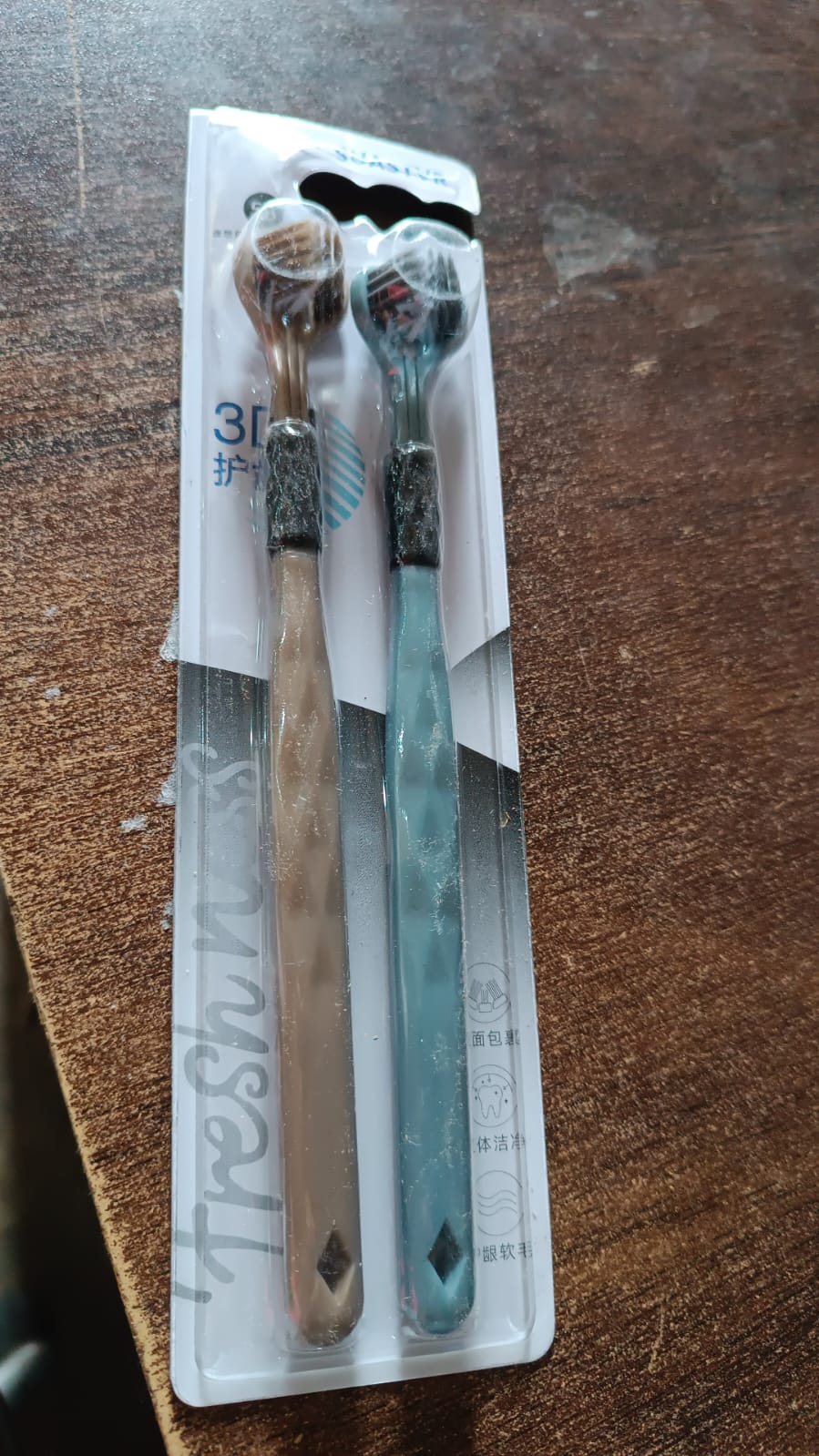 Two three-sided toothbrushes, one beige and one blue, in clear retail packaging on a wooden surface.