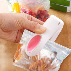 A hand uses a white and pink mini heat sealer to seal a clear bag of snacks on a wooden surface.