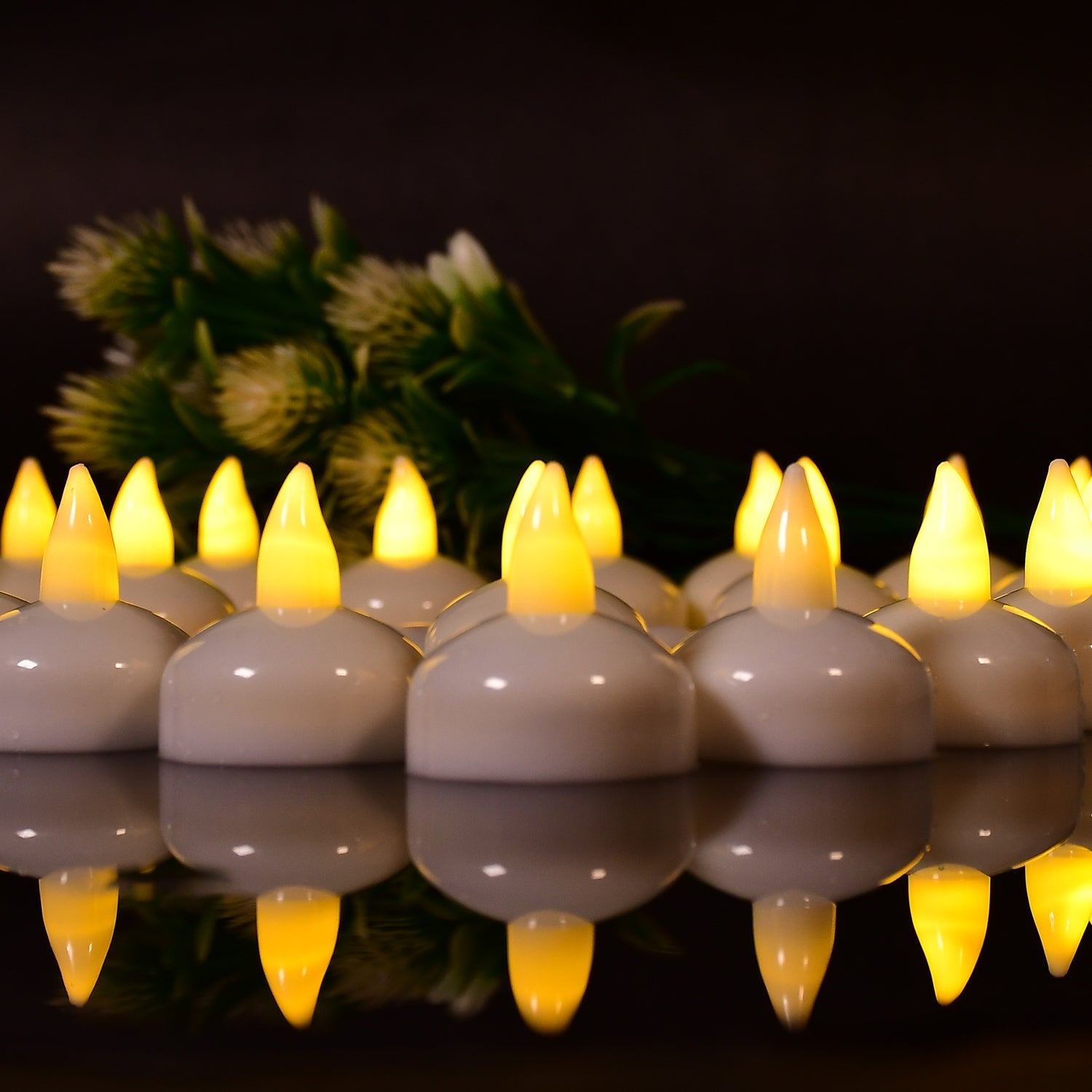 A close-up of multiple white LED floating candles with yellow flames, reflected on a dark surface, with green foliage behind.