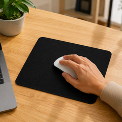 A hand uses a white mouse on a black mouse pad placed on a wooden desk next to a laptop and plant.
