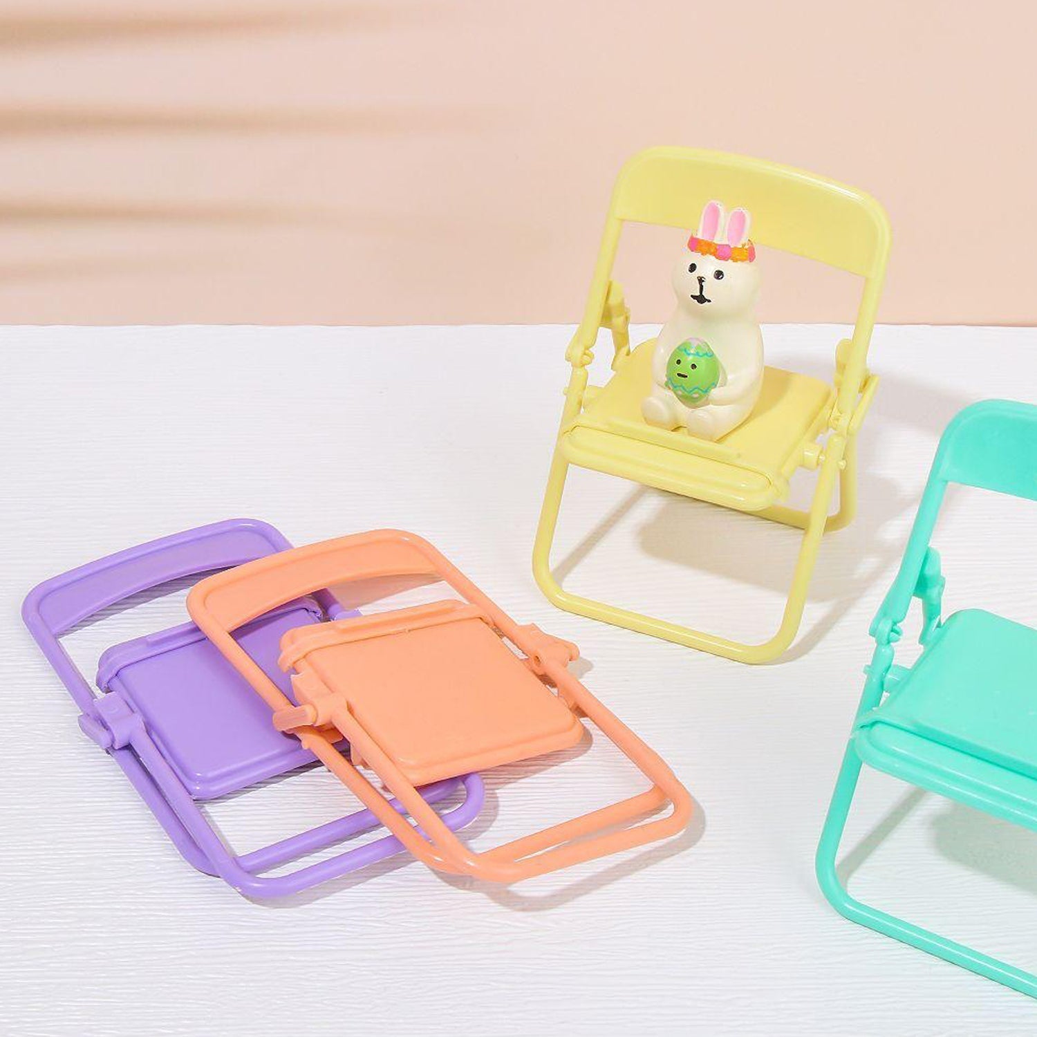 A yellow miniature chair holds a rabbit figurine, while purple, peach, and teal folded chairs are scattered on a surface.