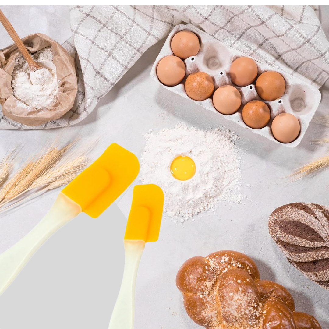 Two yellow silicone spatulas are on a light surface with flour, an egg yolk, a carton of eggs, and baked goods.