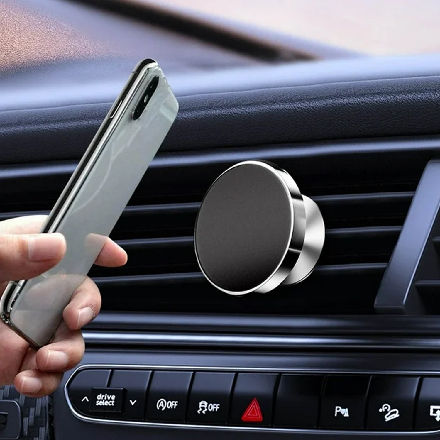 A silver smartphone is held by a hand next to a black and silver magnetic car mount attached to an air vent.