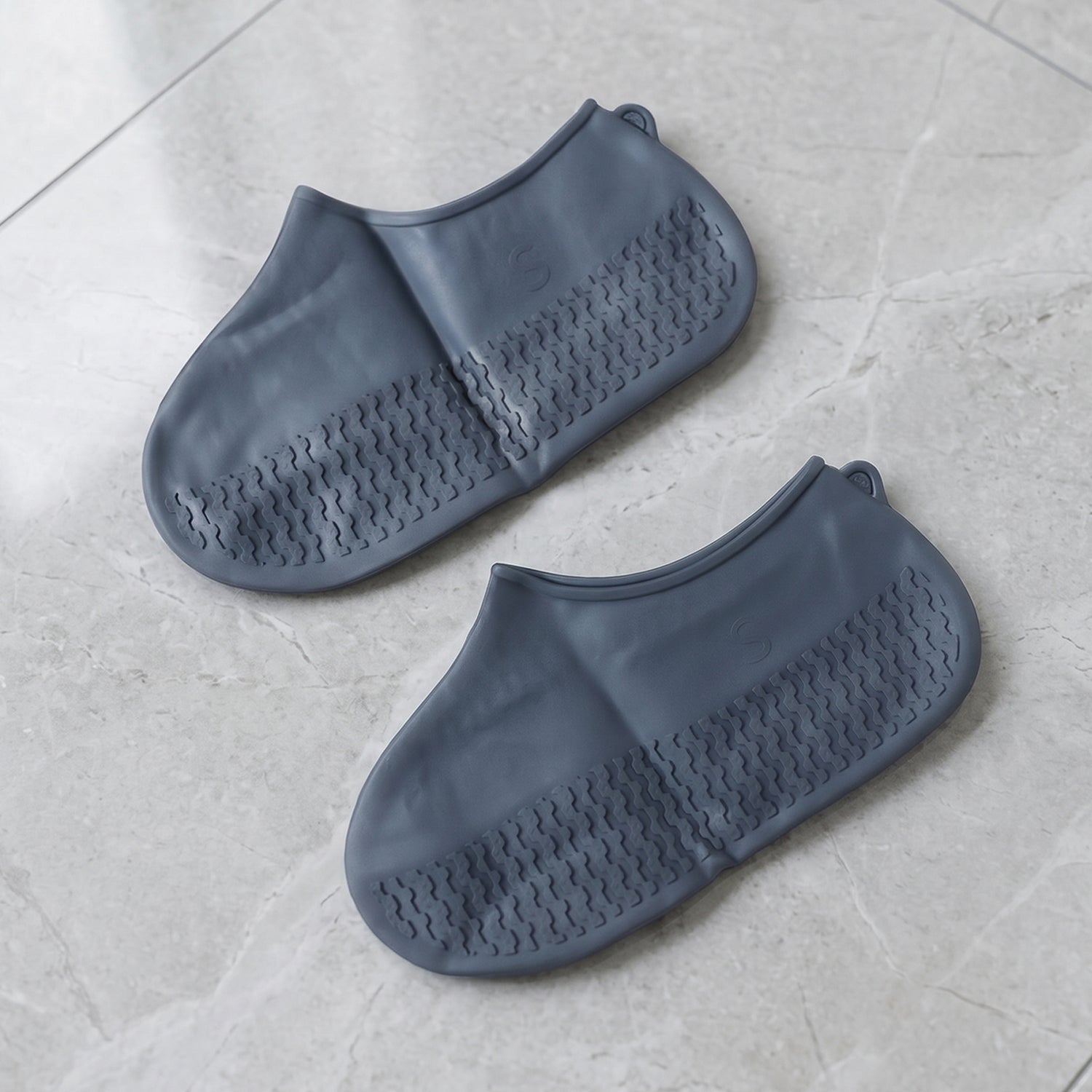 Two dark grey silicone shoe covers with textured soles are neatly placed on a light grey tiled floor.