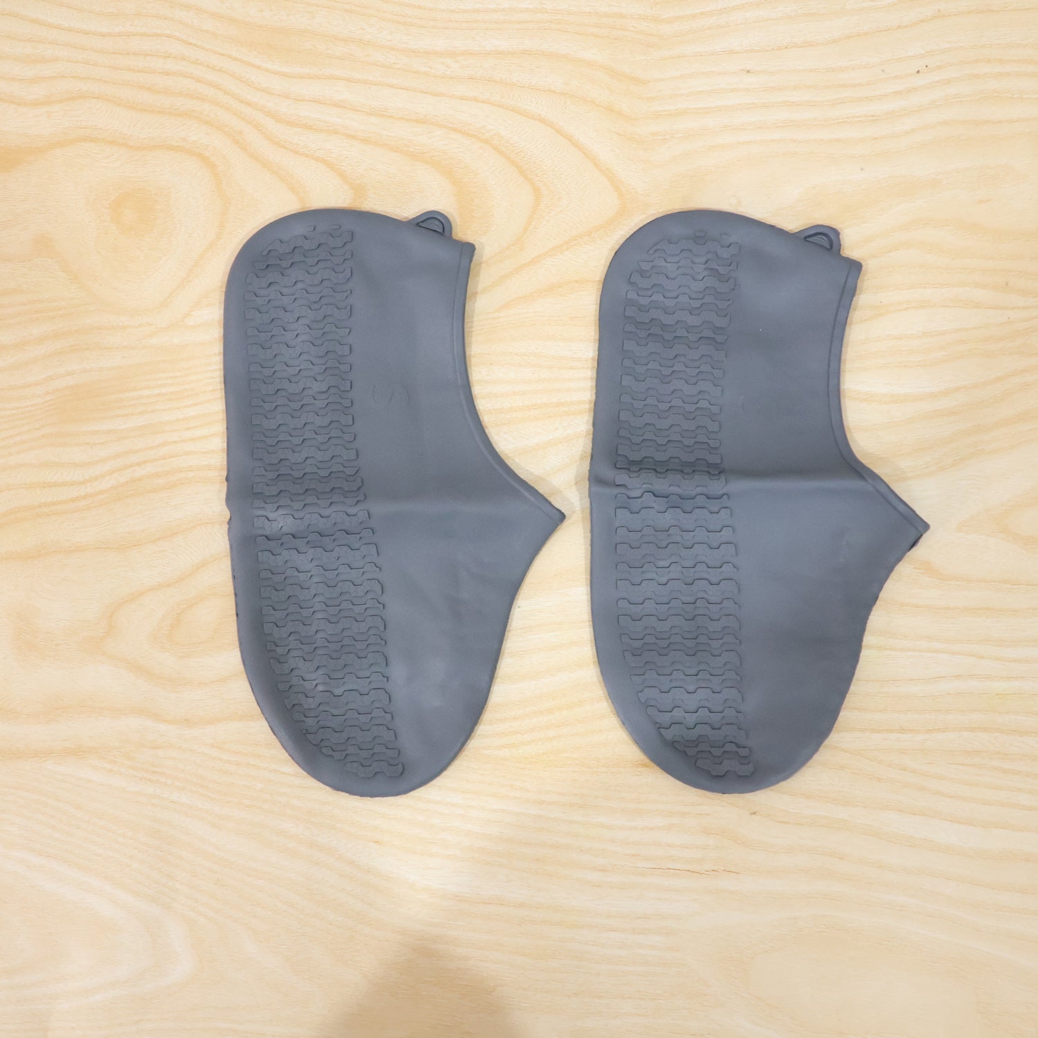 Two dark grey silicone shoe covers with textured soles are laid flat on a light wooden surface.