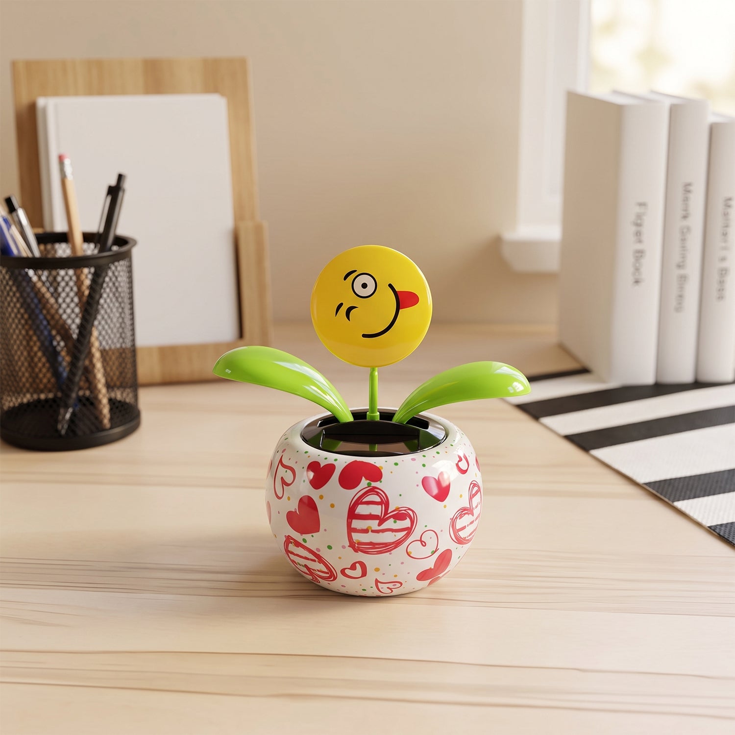 A winking emoji face solar dancing flower in a heart-patterned pot on a light wooden desk next to office supplies.