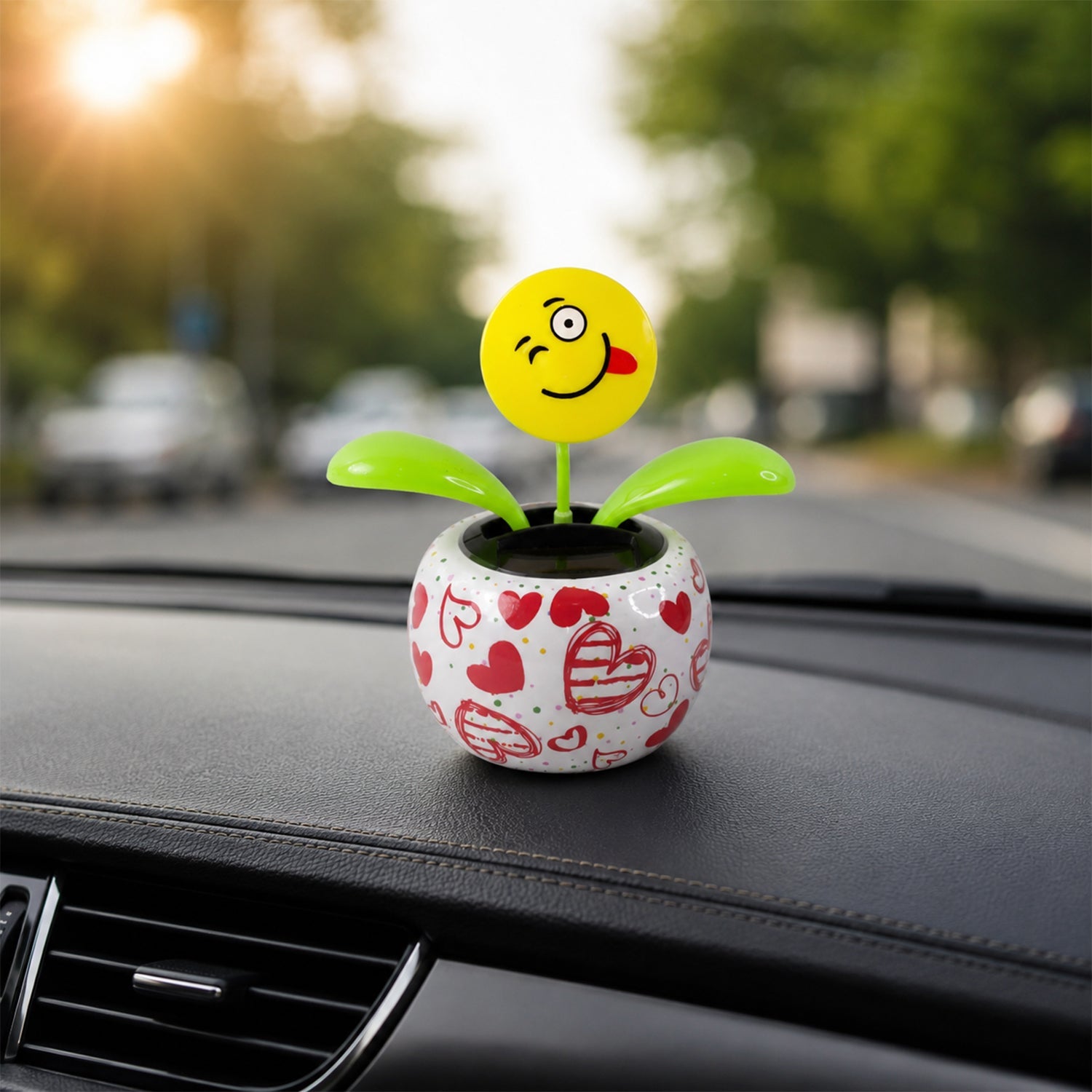 A winking emoji face solar dancing flower in a heart-patterned pot on a car dashboard with sunlight in the background.