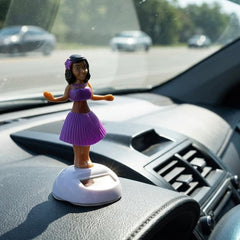 A solar-powered dancing hula girl toy in purple and brown, with a white base, on a car dashboard with cars in the background.