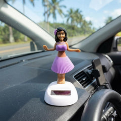 A solar-powered dancing hula girl toy in purple and brown, with a white base, on a car dashboard with palm trees visible.