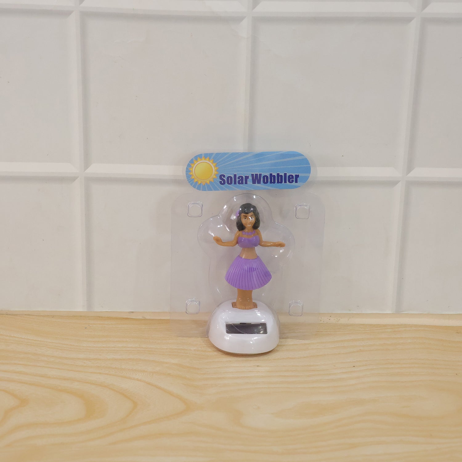 A solar-powered dancing hula girl toy in purple and brown, with a white base, in its clear plastic packaging.