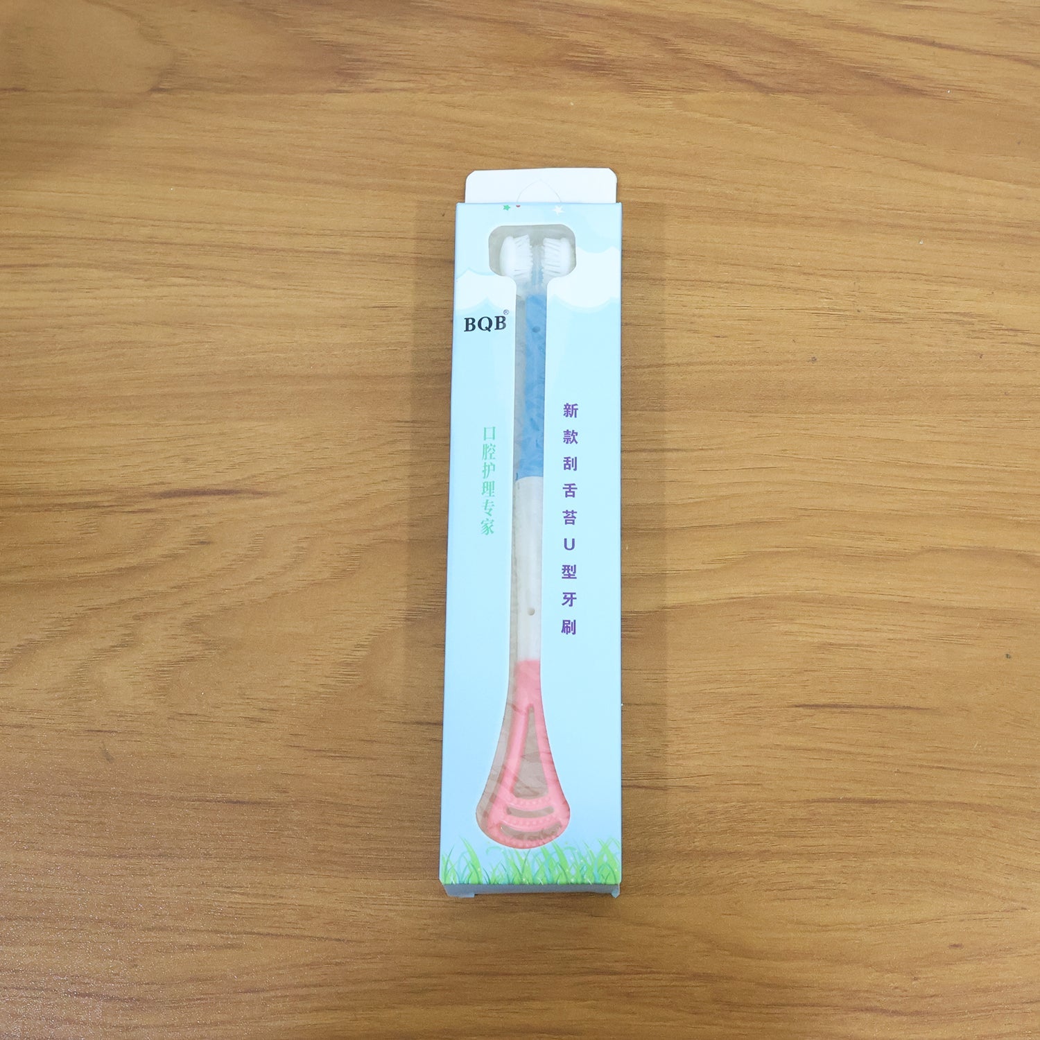 The baby tongue cleaner and toothbrush in its light blue and green packaging, lying on a wooden surface.