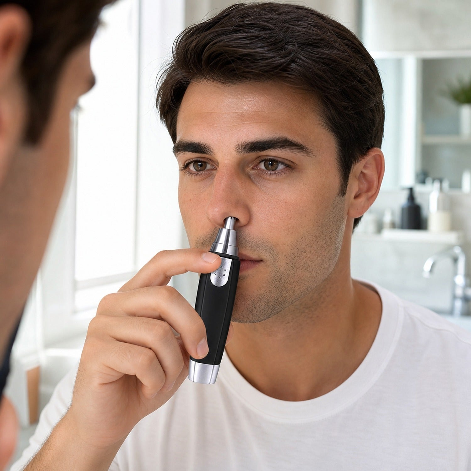 A man with dark hair looks in a mirror while trimming his nose hair with a black and silver device.