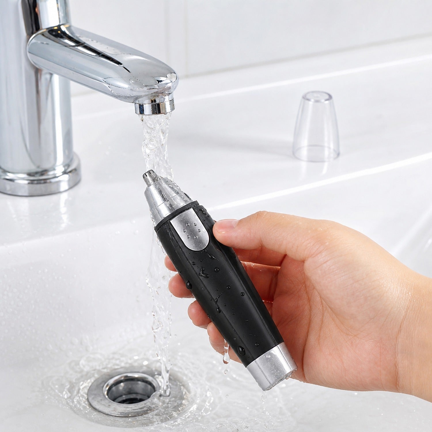 A hand holds a black and silver nose hair trimmer under running water in a white sink.