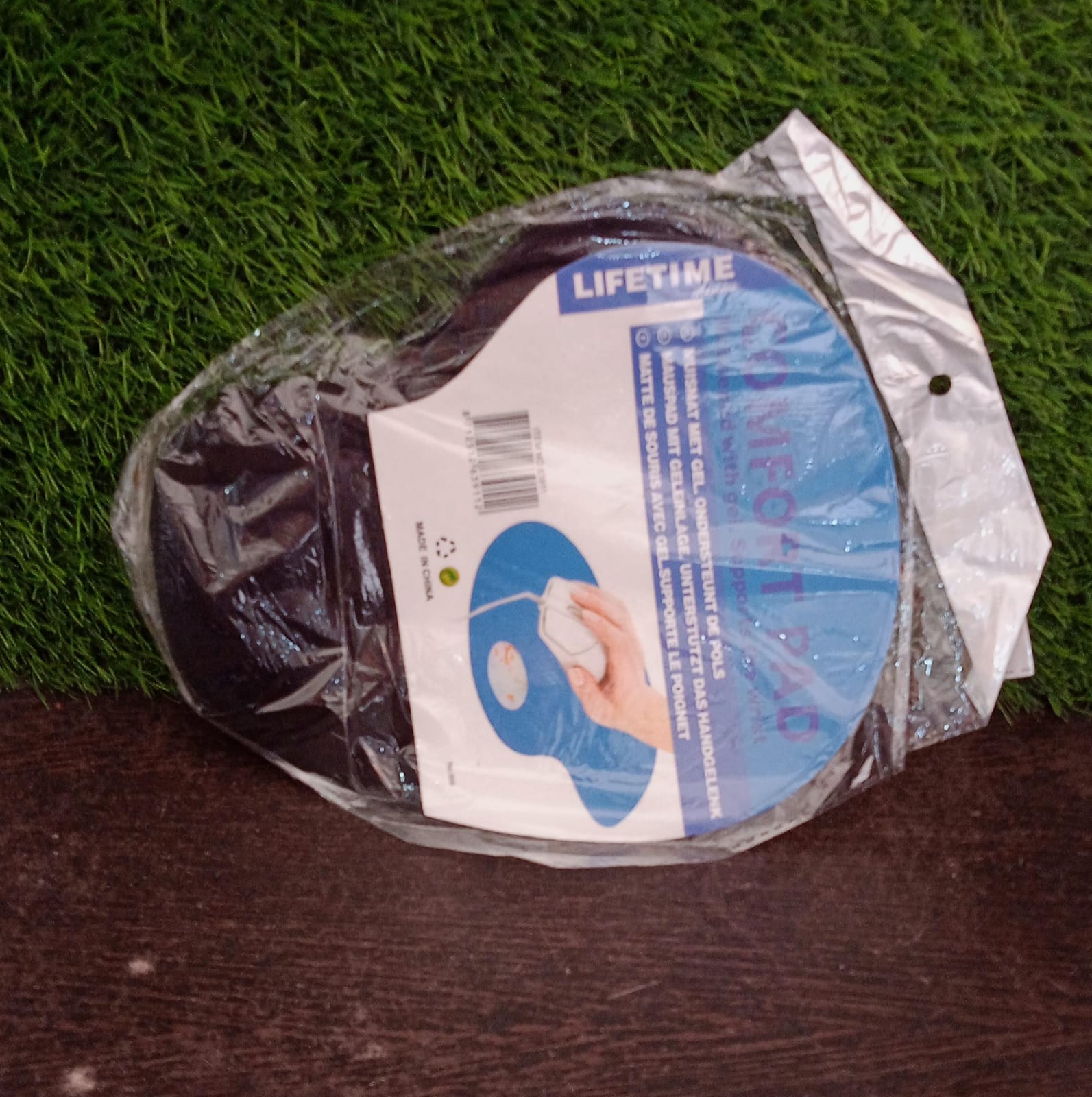 The black ergonomic mouse pad is packaged in clear plastic with a blue label, on a green and brown surface.