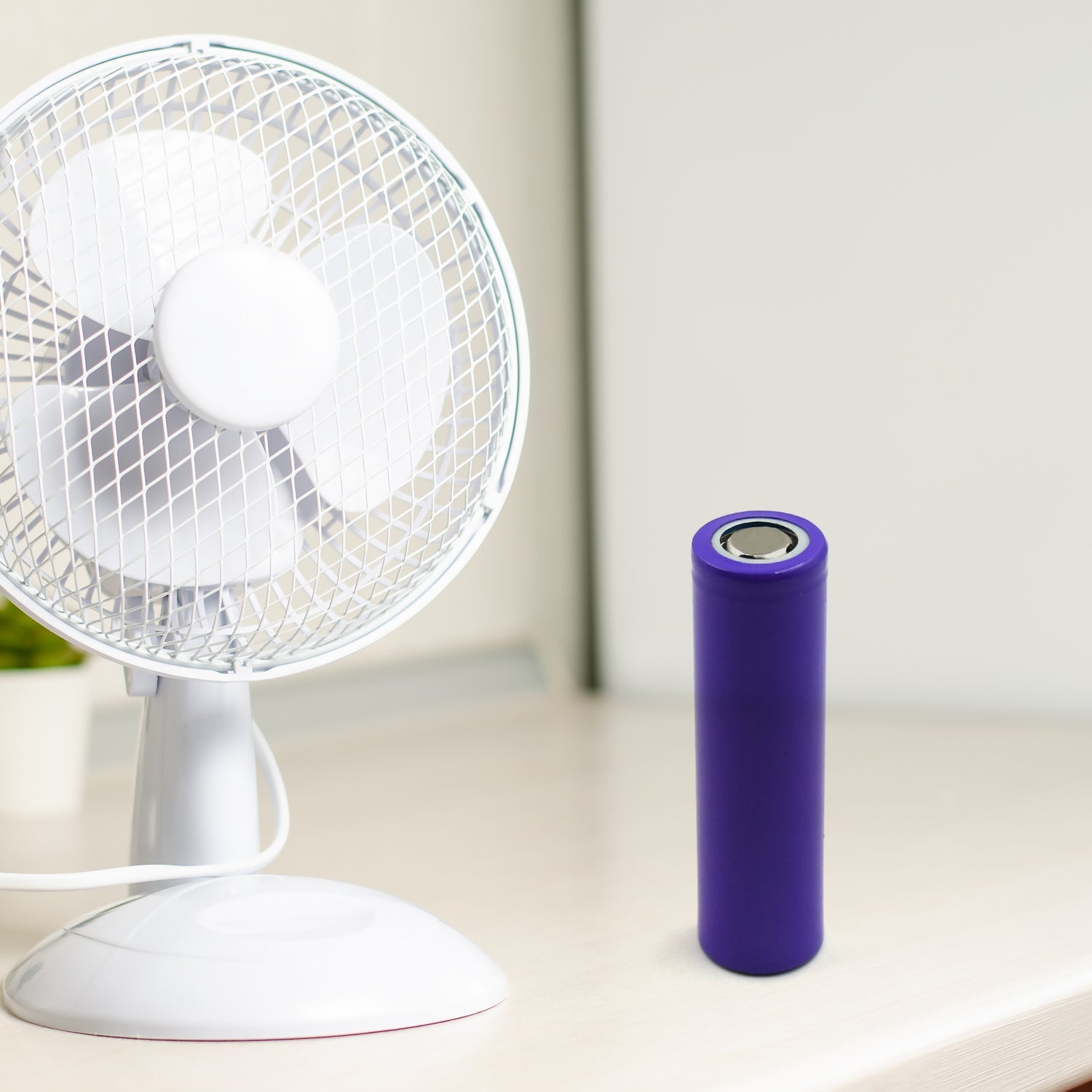 A purple battery stands next to a white desk fan on a light-colored table.